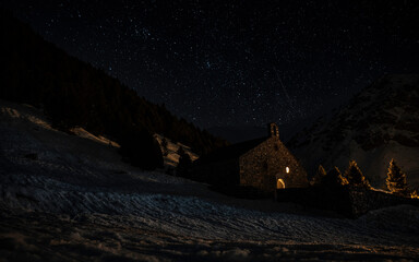 Panoramic night view of the starry sky, Milky Way, and an illuminated stone building on the snow in the foreground.