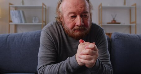 Unhappy elderly man in glasses sitting alone on sofa with clasped hands, looking down in silence. Close up portrait of thoughtful senior male overwhelmed by grief, loneliness and painful memories. - Powered by Adobe