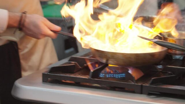 Professional chef working in a commercial kitchen preparing a meal with steam rising from the cooking pans and other kitchen equipment visible The chef is focused on the cooking process, pouring sauce