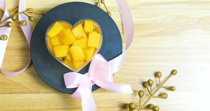 Ripe mango chunks forming heart shape, sitting in golden rimmed dessert container, decorated with pink ribbon on dark plate, presenting romantic valentines dessert