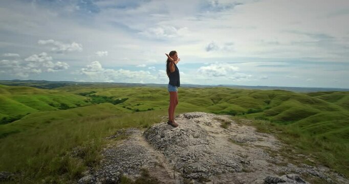 Back view of a young woman with open arms celebrating freedom and enjoying the beautiful landscape on top of green hills in Sumba Island, Indonesia