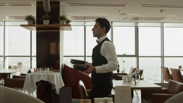 Tracking slowmo shot of young Caucasian waiter in elegant classic outfit bringing ordered dishes in bowls working in fine dining restaurant with large panoramic windows