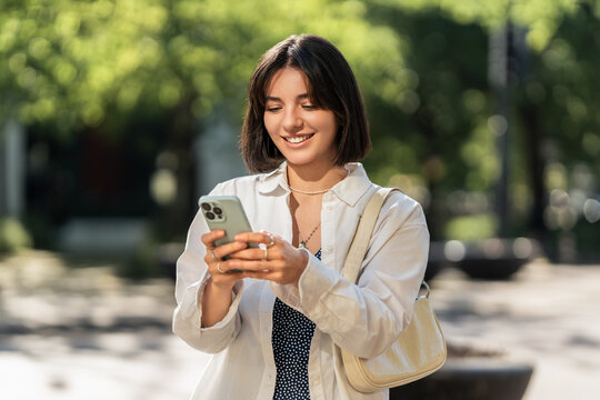 A summer day out. A smiling woman checking an app on her phone in the city. Browsing online shops, chatting with friends and family or checking the map for navigation