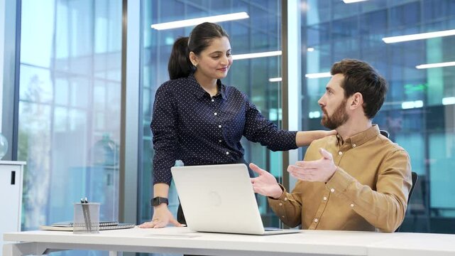 Young woman sexually harasses a male coworker places hand on shoulder at workplace in business office. Beautiful female worker flirts with a shocked and concerned male colleague who is sitting at desk