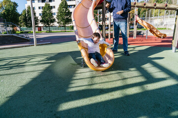 Joyful child sliding down playground slide with caregiver