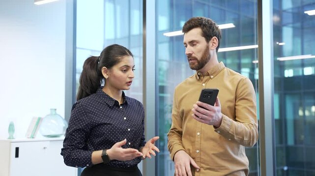 Businessman explains to a female colleague how to use a new application on a mobile phone, talks and teaches. Two coworkers, a man and a woman in office discussing mobile app usage with smartphone