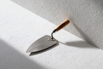 Construction trowel with a wooden handle resting against a textured white wall, casting a dark shadow.