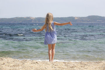 Fototapeta premium Cheerful little girl in blue summer dress dancing on the beach. Concept of freedom, happiness and positivity 