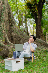 Fototapeta premium Young man working on laptop in serene park atmosphere surrounded by large trees during a sunny day