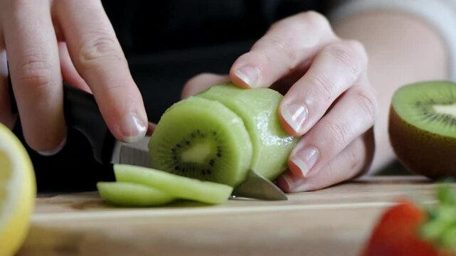 Hand cuts peeled kiwi into thin slices surrounded by other fresh fruits