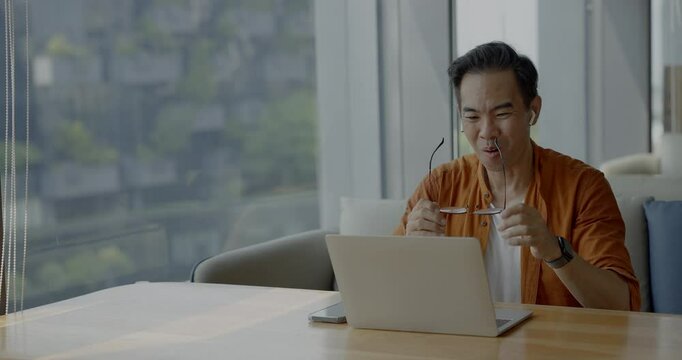 Senior Asian businessman wearing headset celebrates success during online meeting in modern co-working space. Remote work and business communication concept.