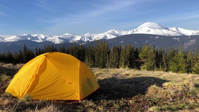 Tent on foreground in the mountains, first morning light. Campsite with a yellow dome tent on a high mountain bluff on the background of snowy mountains at sunrise