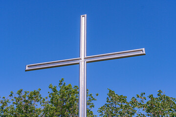 A large metal cross stands against a clear blue sky, possibly in a park or natural setting, with lush green trees and shrubbery in the background The composition draws attention upward from the grou