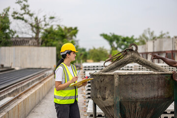 Female worker or engineer wearing safety gear, a yellow hard hat and vest. Construction worker is holding a clipboard standing near a large concrete bucket as part of an active construction operation.