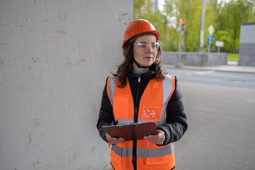 A female civil engineer in an orange construction helmet and vest stands under an overpass and...