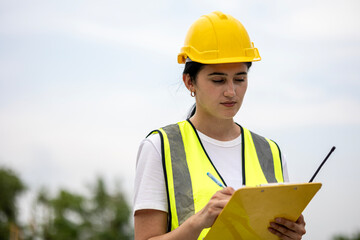 Female worker or engineer wearing safety gear, a yellow hard hat and vest. Construction worker is...