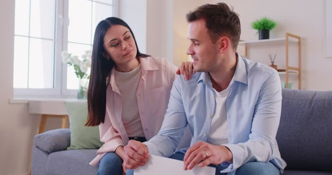 Unhappy young man in worry, wife supporting, couple sits to solve, separate problems in relationship, thinking, financial paper troubled, worried young spouses deal with debt bankruptcy, business