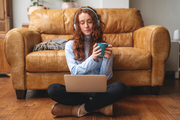 Young woman enjoys coffee and music while working on laptop in cozy living room with leather couch...