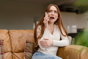 Woman on the phone expressing concern while sitting on a couch in a modern living room during...