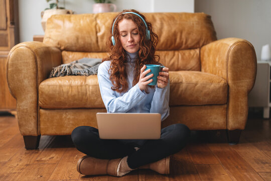 Young woman enjoys coffee and music while working on laptop in cozy living room with leather couch during relaxed afternoon hours