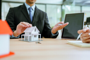Asian real estate agent in modern office consulting clients, showcasing properties with a laptop, tablet, and house model.