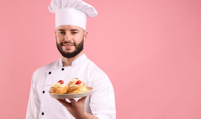 Happy confectioner in uniform holding profiteroles with strawberries on pink background