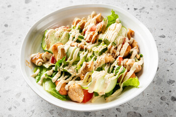 Fresh salad with grilled chicken, lettuce, cucumber, tomatoes, sesame seeds, and creamy dressing in a white bowl on a speckled countertop. Healthy, nutritious, and appetizing meal.