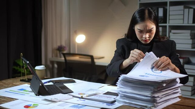 A woman is sitting at a desk with a laptop and stacks of papers. She is working on a project or report