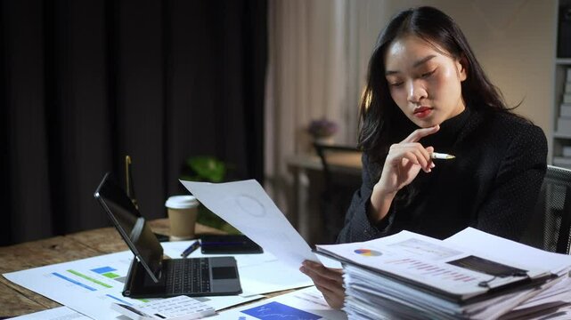 A woman is sitting at a desk with a laptop and a stack of papers. She is writing on a piece of paper with a pen