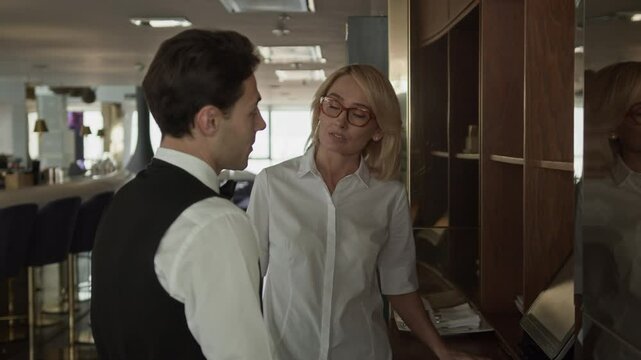 Medium shot of middle-aged Caucasian restaurant manager engaged in conversation with young waiter in elegant classic outfit standing near computer screen of electronic POS system in fancy restaurant
