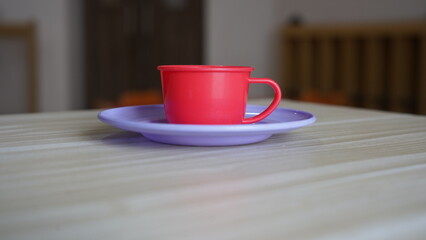 A red plastic toy teacup sits on a lavender plastic toy saucer on a light-colored table