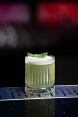 A pale yellow cocktail with a frothy white top is garnished with two vibrant green mint leaves, sitting on a reflective bar surface.