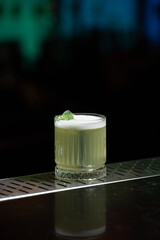 A pale yellow cocktail with a frothy white top is garnished with two vibrant green mint leaves, sitting on a reflective bar surface.