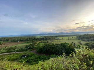 Peaceful rural scenery at sunrise with green rice fields, trees, hills and mountains in the distance under colorful sky in Garut West Java, Indonesia