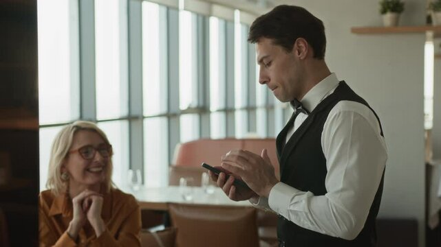 Medium side view shot of young Caucasian waiter in classic outfit using gadget while taking order from elegant middle-aged woman and her unrecognizable guest sitting at dining table in restaurant