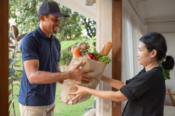 African American male Delivery smiles while handing a bag filled with fresh groceries to Asian...