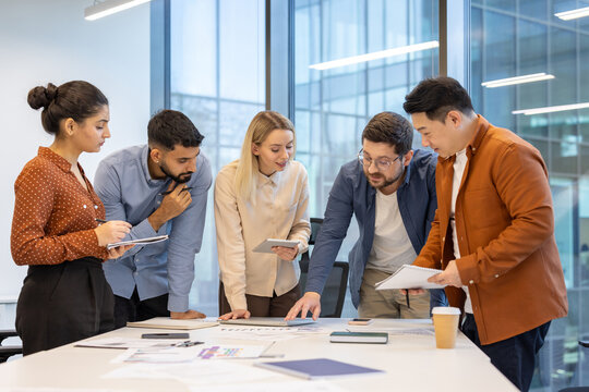 A diverse group of professionals collaborate around a table, reviewing documents and planning strategies in the office setting.