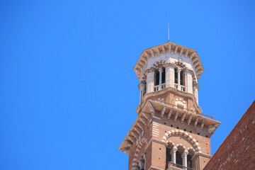 The historic tower rises prominently against a clear blue sky in Verona, Italy. Its striking architecture features intricate stonework and distinct levels, showcasing the city's rich heritage.