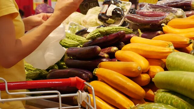 A shopper inspects a bitter melon, turning it over thoughtfully. The scene captures a natural moment of selection with vibrant tropical vegetables in the background.