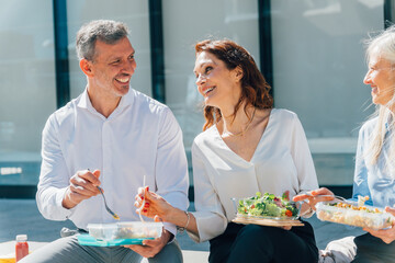 Business professionals sharing nutritious lunch outdoors, smiling and conversing, highlighting workplace collaboration and well-being during midday break