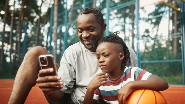 African American child talking and spending time together with his dad at basketball court. Boy with father posing for smartphone camera while taking selfie after playing basketball outdoors. - Powered by Adobe