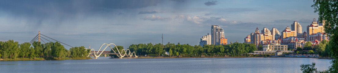Modern residential buildings and pedestrian bridge on the Dnipro River in Kyiv at sunset. Peaceful cityscape with calm water, trees, and dramatic sky.