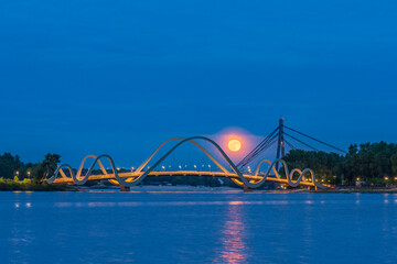 Full moon rises over the Dnipro River in Kyiv, glowing behind a modern pedestrian bridge. City lights and calm water create a peaceful and dreamy evening atmosphere.
