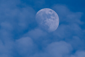 Close-up of the waxing gibbous moon in the blue daytime sky with soft clouds. A rare and peaceful view of the moon clearly visible before nightfall.