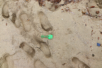 A small green plastic shovel is partially buried in sand on a beach. Surrounding it are multiple footprints, suggesting recent activity. The sand is a light beige color.