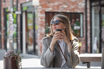 Beautiful woman in sunglasses holding disposable cup of coffee while sitting on open veranda in cafe