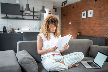 Focused freelancer reading documents on sofa with laptop in home office