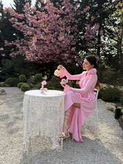 beautiful woman with dark hair in luxurious pink dress and accessories drinking tea in blooming...