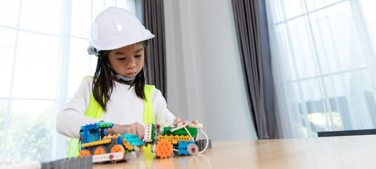 A young girl wearing a safety helmet and reflective vest is playing with engineering toys,...