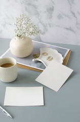 Blank paper sheet cards on home office desk workspace with coffee cup, vase, home decor on grey background. Wedding invitation cards. Flat lay, top view mockup with empty copy space.Life style concept